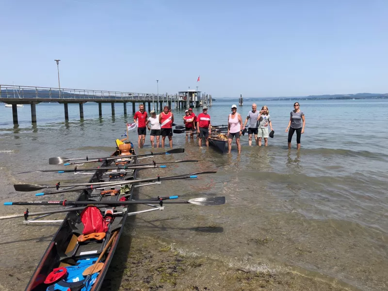 FRGS-Ruderinnen und -Ruderer mit ihren Booten am flachen Bodensee-Ufer, Steg mit Flagge im Hintergrund
